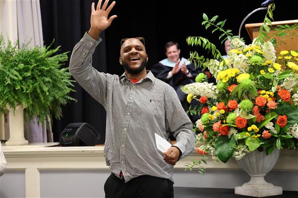 An alum from the Helen Keller School of Alabama waves while smiling broadly and wearing a button up shirt and dress pants. In his left hand, he holds an envelope. 